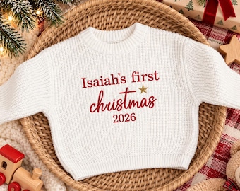 Puede incluir: Un suéter de punto blanco de manga larga con texto bordado en rojo que dice Isaiah's first christmas 2026 y un pequeño acento de estrella dorada sobre la palabra christmas, sobre una cesta redonda.
