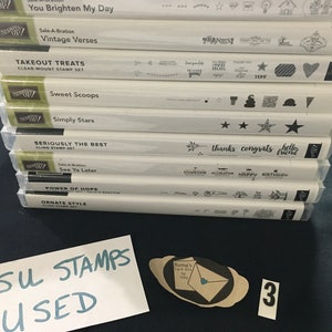 May include: A stack of ten white boxes with black text, labeled with different names of stamp sets. The boxes are stacked on a dark blue surface. A white card with the text "SU STAMPS USED" is in the lower left corner of the image. A small brown card with a white envelope and the text "Roma's Card Kiss by YOU" is in the lower right corner of the image. The number "3" is written on a white piece of paper in the lower right corner of the image.