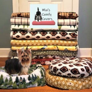 May include: A stack of patterned fleece blankets in various designs and colors, including geometric, floral, and nature-themed prints. A small dog bed with a forest print and a round dog bed are in the foreground. The image includes the text "Mia's Comfy Covers".