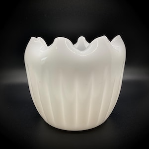 May include: A white ceramic planter with a scalloped rim and vertical ribbed detailing. The glossy finish reflects light, highlighting the curves of the vessel. The planter is set against a dark background.