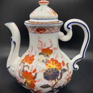 May include: White ceramic teapot with a lid, featuring orange, blue, and gold floral designs. The handle and spout are white with blue accents. The teapot has a bulbous body and a curved handle.