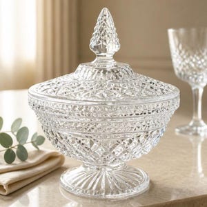 May include: A clear glass candy dish with a lid, featuring a detailed diamond-cut pattern across the bowl and cover. The lid is topped with a pointed finial, and the entire piece rests on a circular, fluted pedestal base for stability.
