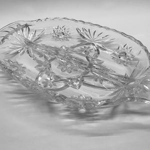 May include: Clear, oval-shaped glass serving dish with starburst and floral designs. The dish is divided into three sections and features a scalloped edge and handles. A decorative piece for serving appetizers or desserts.