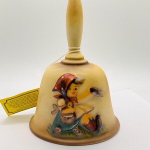 May include: A vintage bell with a cream-colored handle and a bell-shaped body. The bell features a painted scene of a girl with a red headscarf, a basket, and a duckling. The bell is a collectible item.