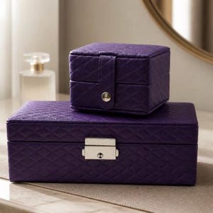 May include: Two purple jewelry boxes with a quilted texture. The larger rectangular box features a silver-toned metal latch closure, while the smaller square box has a snap button closure. Both boxes are constructed with a consistent diamond pattern.