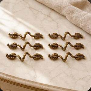 May include: A set of six antique brass-toned metal drawer pulls, each featuring a winding, vine-like stem design with a single leaf at each end. The hardware pieces are arranged in two columns of three, showcasing a metallic finish.
