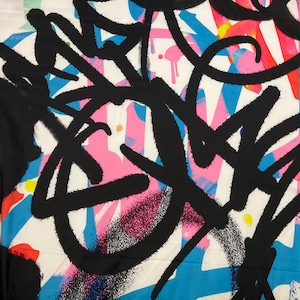 May include: A white fabric featuring a vibrant graffiti-style print with bold black calligraphic lines layered over splashes of bright blue, hot pink, and red paint. The design includes scattered small yellow and orange dots throughout the pattern.