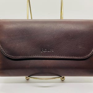 May include: A dark brown leather wallet with a flap closure. The wallet has the name "Rolfs" embossed on the front. The wallet is displayed on a gold-colored stand against a white background.
