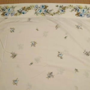 May include: White bed sheet with a floral pattern. The top edge features a border of blue roses and green leaves. Scattered across the sheet are smaller clusters of blue and green flowers. The overall design is soft and delicate.