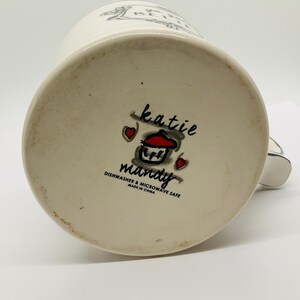 May include: A white ceramic mug with a black handle, featuring the text "BE HAPPY" and a decorative design. The bottom of the mug displays the "katie mandy" logo with a cupcake illustration and the text "Dishwasher & Microwave Safe Made in China."