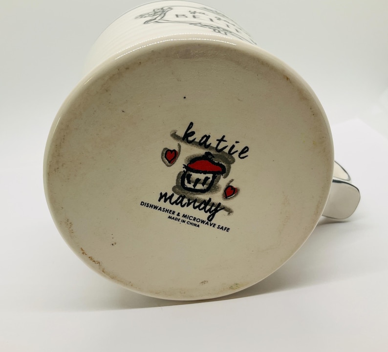 May include: A white ceramic mug with a black handle, featuring the text "BE HAPPY" and a decorative design. The bottom of the mug displays the "katie mandy" logo with a cupcake illustration and the text "Dishwasher & Microwave Safe Made in China."