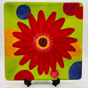 May include: Square decorative plate with a bright red flower design on a lime green background. The flower has a yellow center with a black dot, and the plate is decorated with yellow, red, and blue polka dots.