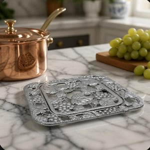 May include: A square silver-toned metal trivet featuring an intricate embossed pattern of grape clusters and vine leaves. The trivet has a flat, openwork design with a raised border, providing a stable surface for hot cookware items.