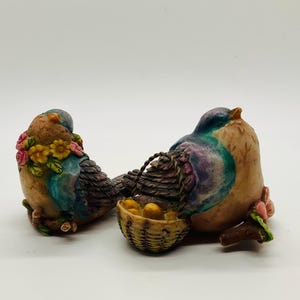 May include: Two decorative bird figurines. One bird has a floral wreath and the other holds a small basket with yellow objects. The birds are painted in shades of blue, brown, and purple, with a touch of pink.