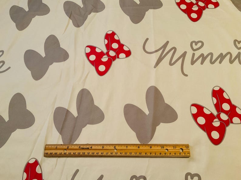 May include: White fabric with a repeating pattern of grey bows and red bows with white polka dots. The text "Minnie" is printed in grey on the fabric.
