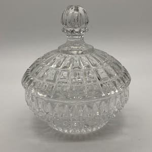 May include: A clear, faceted glass candy dish with a lid. The dish has a round base and a domed lid with a knob handle. The glass has a diamond-like pattern, reflecting light. A decorative piece for storing small items.