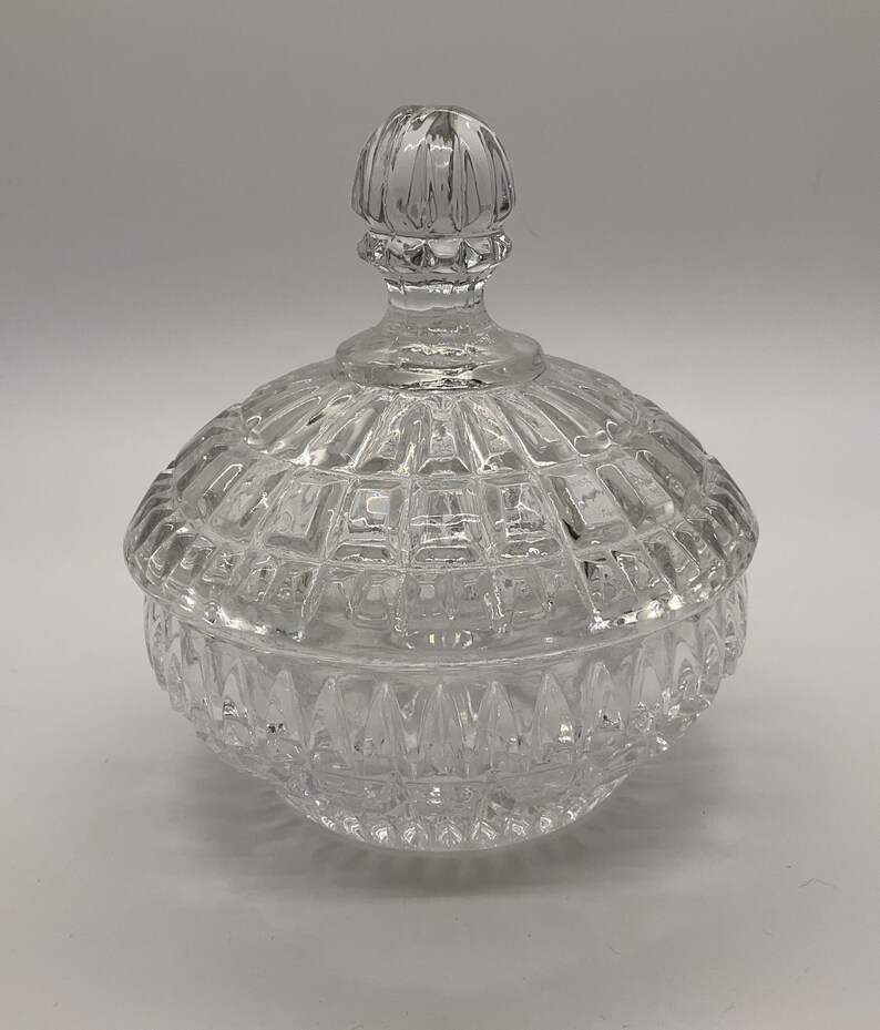 May include: A clear, faceted glass candy dish with a lid. The dish has a round base and a domed lid with a knob handle. The glass has a diamond-like pattern, reflecting light. A decorative piece for storing small items.