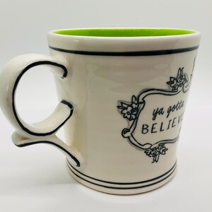 May include: Cream-colored ceramic mug with a green interior. The mug features a black-lined handle and rim, with the words "ya gotta BELIEVE" in a decorative frame. Floral accents surround the text.