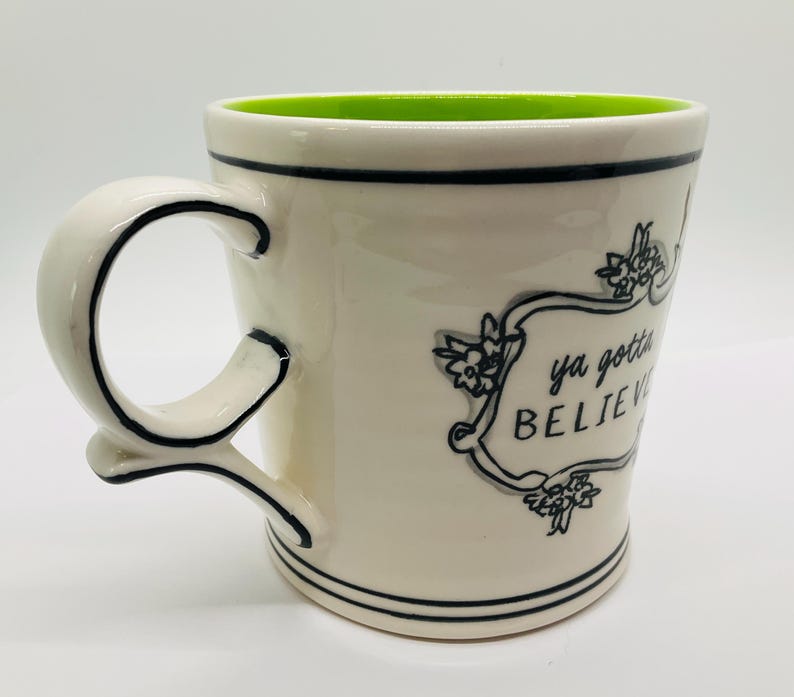 May include: Cream-colored ceramic mug with a green interior. The mug features a black-lined handle and rim, with the words "ya gotta BELIEVE" in a decorative frame. Floral accents surround the text.