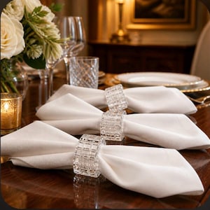 May include: Three white fabric napkins folded and secured with clear, square-cut crystal napkin rings. The rings feature a grid-like, faceted texture that reflects light, holding each napkin in a neat, uniform shape on a dark wooden surface.