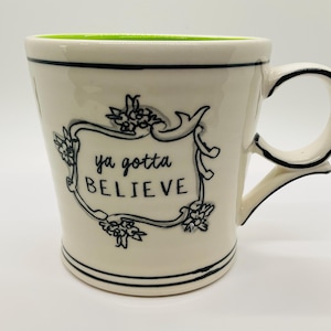May include: Cream-colored ceramic mug with a black floral design and the words "ya gotta BELIEVE" in a decorative frame. The mug has a black handle and trim, and a bright green interior.