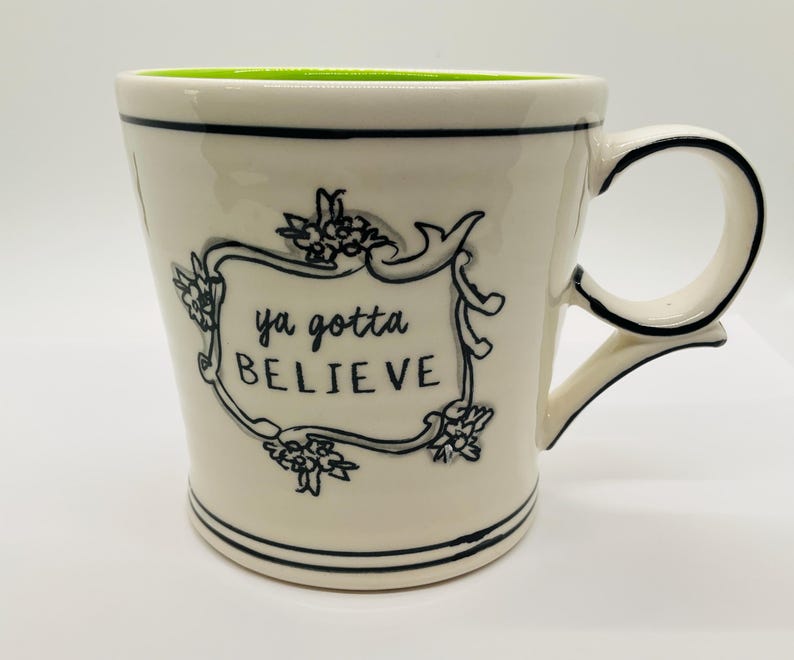 May include: Cream-colored ceramic mug with a black floral design and the words "ya gotta BELIEVE" in a decorative frame. The mug has a black handle and trim, and a bright green interior.