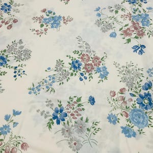 May include: A white fabric featuring a repeating floral pattern. The design consists of scattered bouquets of small flowers in shades of blue, dusty pink, and grey, accented with green leaves and stems distributed evenly across the surface.