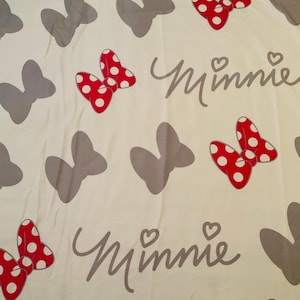 May include: White fabric with a repeating pattern of grey bows and red bows with white polka dots. The text "Minnie" is written in grey cursive.