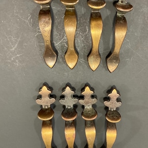May include: Eight antique bronze cabinet pulls. Each pull features a decorative top with a stylized design, a curved handle, and a pointed base. The pulls have a warm, aged patina, suggesting they are vintage or vintage-inspired.