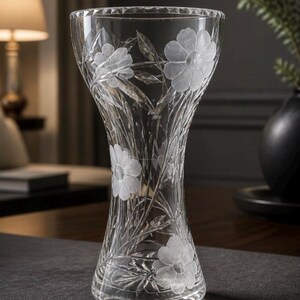 May include: A clear glass vase with a flared rim and a tapered waist, featuring etched floral patterns of daisies and slender leaves. The glass has a faceted, diamond-cut texture along the top edge and a smooth, transparent body with frosted details.