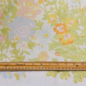May include: Floral fabric with a vintage design. The fabric features large flowers in shades of pink, yellow, and orange, with green leaves and stems on a white background. A wooden ruler is placed across the fabric, measuring up to 12 inches.
