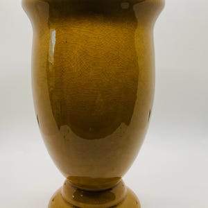 May include: A tall, glazed ceramic vase in a warm, mustard yellow hue. The vase has a wide, flared top and a pedestal base. The surface displays a crackle pattern, adding texture. This decorative vase is suitable for floral arrangements or as a standalone accent piece.