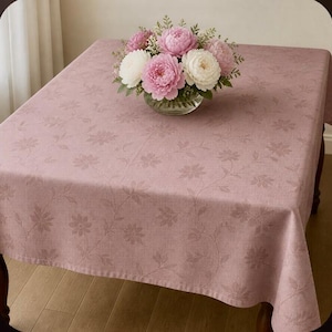 May include: A dusty rose tablecloth featuring a subtle, tonal floral pattern across its surface. A glass vase sits in the center, filled with a bouquet of artificial pink and white peony flowers with green leaves and stems.
