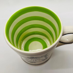 May include: White ceramic mug with a green and white striped interior. The mug has a curved handle with a black outline. The exterior features a floral design and text. A decorative kitchenware item.