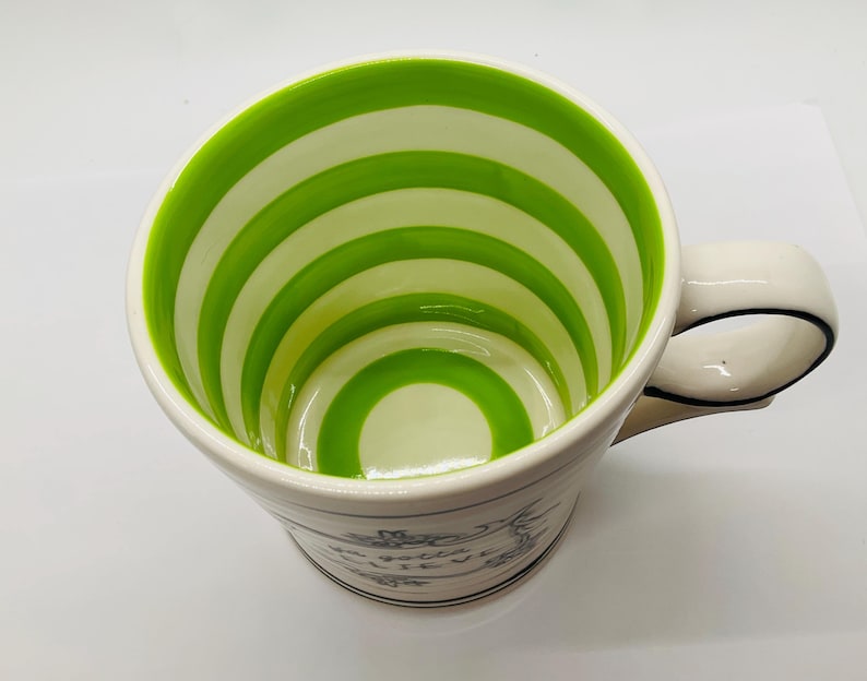 May include: White ceramic mug with a green and white striped interior. The mug has a curved handle with a black outline. The exterior features a floral design and text. A decorative kitchenware item.