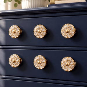 May include: A dark navy blue dresser featuring six circular drawer knobs. Each knob is crafted with a gold-toned metal base and a white floral-shaped inlay, showcasing intricate petal details and a small gold center point on the front surface.