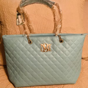 May include: A light blue quilted tote bag with gold-colored chain handles. The bag features a gold-colored logo on the front and a diamond pattern. The tote bag is a stylish accessory.