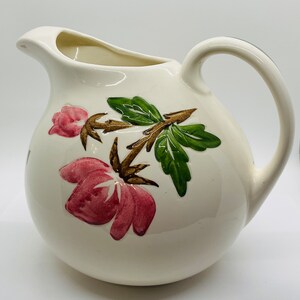 May include: White ceramic pitcher with a floral design. The pitcher has a round body and a handle. The floral design features pink flowers, green leaves, and brown stems.