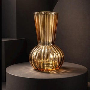 May include: A golden-colored glass vase with a bulbous base and a fluted, cylindrical neck. The vase sits on a dark gray circular pedestal. The glass has vertical ridges, creating a textured surface. The overall design is elegant and decorative.