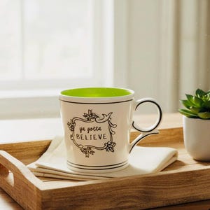 May include: White ceramic mug with a green interior and the words "ya gotta BELIEVE" in a black floral frame. The mug sits on a white napkin on a wooden tray, with a small potted succulent plant nearby.
