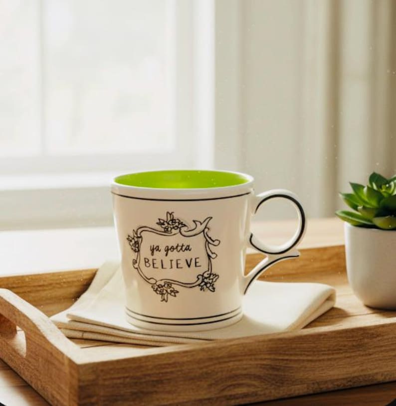 May include: White ceramic mug with a green interior and the words "ya gotta BELIEVE" in a black floral frame. The mug sits on a white napkin on a wooden tray, with a small potted succulent plant nearby.