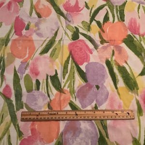 May include: Floral fabric with a watercolor design featuring pink, peach, and purple flowers with green leaves on a white background. A wooden ruler is placed across the bottom of the fabric.