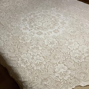 May include: A white lace tablecloth with a floral pattern. The intricate design features a central medallion and a detailed border of swirling patterns and flowers. The delicate fabric is spread across a surface.