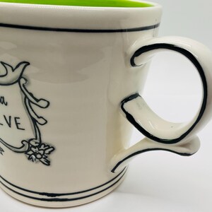 May include: Cream-colored ceramic mug with a green interior and a black handle. The mug features the phrase "gonna believe" in a decorative banner. Black lines accent the rim and base.