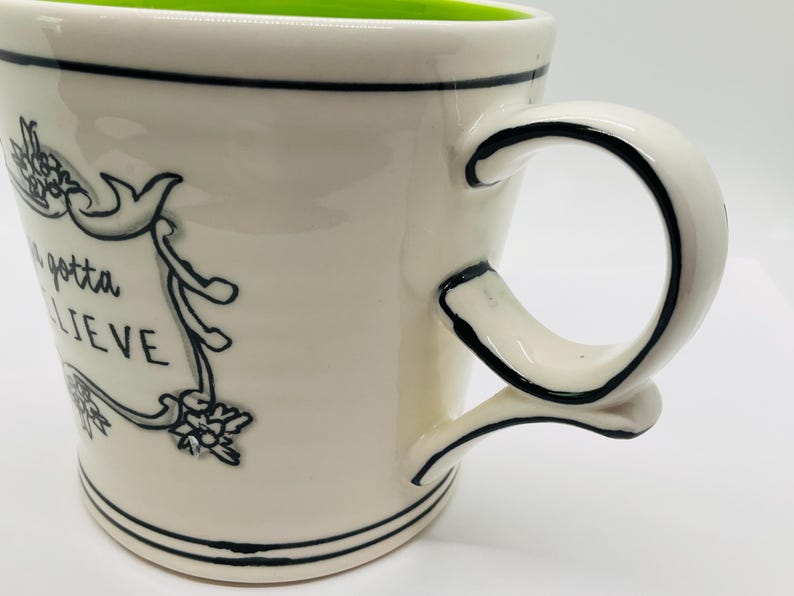 May include: Cream-colored ceramic mug with a green interior and a black handle. The mug features the phrase "gonna believe" in a decorative banner. Black lines accent the rim and base.