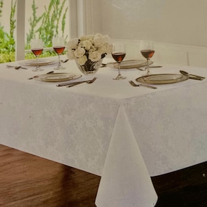 May include: A white tablecloth with a subtle, tone-on-tone floral damask pattern draped over a table. The table is set with four wine glasses filled with red wine, four plates, and silverware. A glass bowl filled with white roses sits in the center.