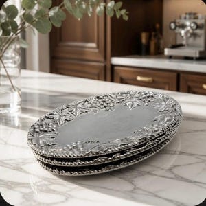 May include: A stack of three oval silver-toned metal platters featuring a raised relief border design of grape clusters and vine leaves. The platters have a smooth, reflective central surface and a textured, rope-like edge detail around the rim.