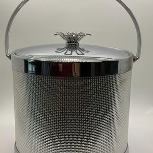 May include: A silver ice bucket with a textured, woven-like pattern. It has a lid with a decorative floral-shaped handle and a curved handle for carrying. The bucket is cylindrical and made of shiny metal.