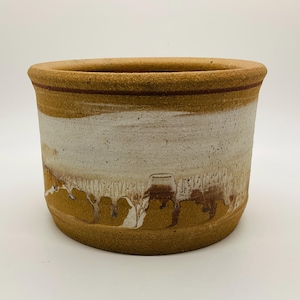 May include: A handcrafted ceramic planter with a cylindrical shape. The planter features a light brown rim with a thin red stripe, a white band, and a lower section with brown and tan dripped glaze. The overall aesthetic is rustic and earthy.