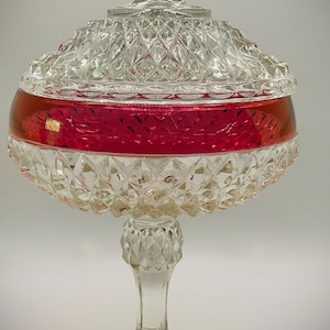 May include: A clear glass candy dish with a diamond pattern and a red band around the middle. The dish has a lid with a pointed finial and sits on a pedestal base. The candy dish is a decorative item.